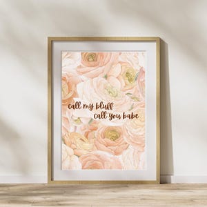 Puede incluir: Una impresión floral enmarcada con un diseño de acuarela de rosas color melocotón y crema. La obra de arte incluye el texto "call my bluff call you babe" en un estilo manuscrito. El marco es de color madera clara.