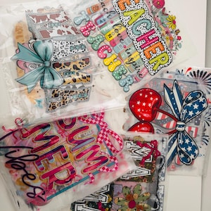 May include: Five clear plastic bags containing iron-on heat transfer vinyl designs. The designs include "Teacher", "Summer Vibes", "Mama", and two patriotic designs with red, white, and blue colours.