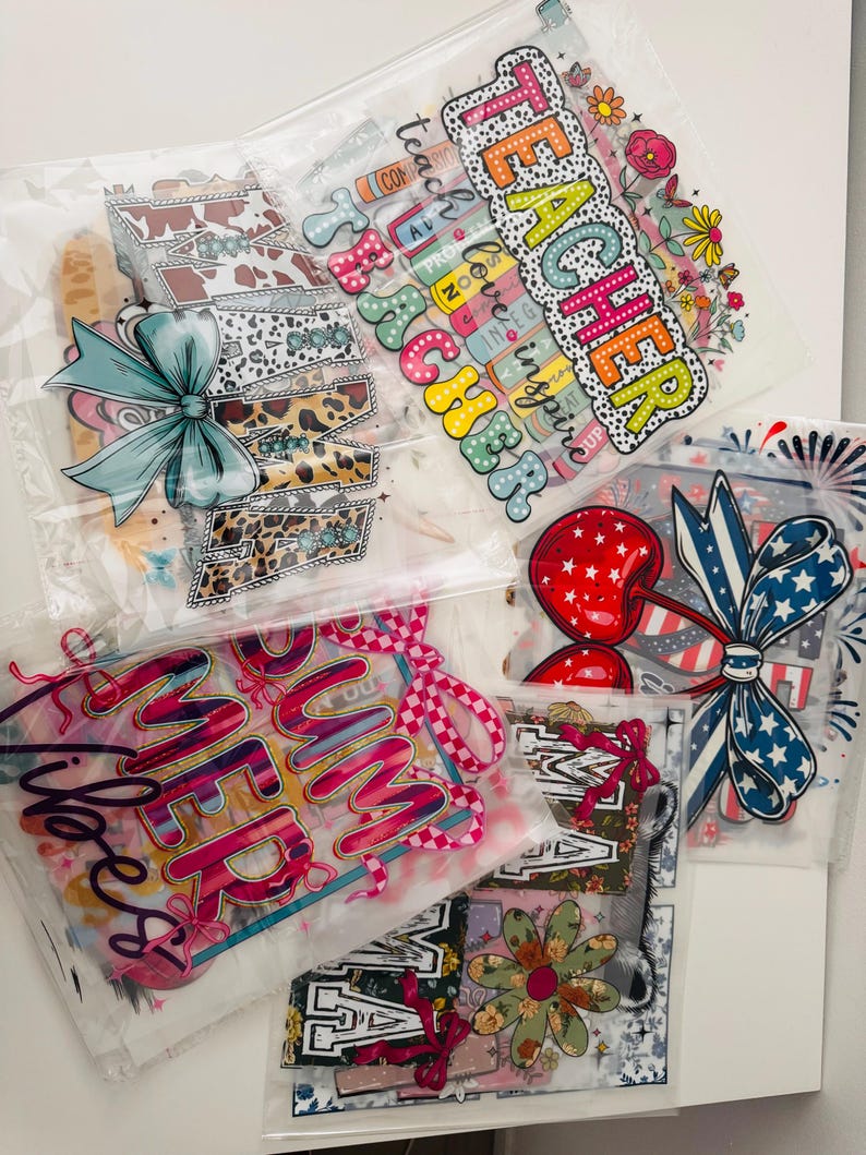May include: Five clear plastic bags containing iron-on heat transfer vinyl designs. The designs include "Teacher", "Summer Vibes", "Mama", and two patriotic designs with red, white, and blue colours.