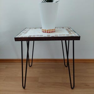 Puede incluir: Una mesa pequeña, cuadrada, de mosaico con un marco de madera oscura y patas de horquilla negras. Un macetero blanco con una planta suculenta verde se encuentra encima de la mesa.