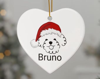 May include: A heart-shaped white ceramic ornament featuring a cartoon dog wearing a Santa hat. The dog's name, "Bruno," is printed in black below the illustration. A gold string is attached for hanging.