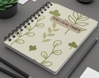 May include: Spiral-bound notebook with a cream-colored cover featuring a repeating pattern of green leaves and clovers. The text "Wild & Unruly Thoughts" is displayed on a rectangular label. Two pencils are visible.