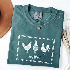 May include: Teal Comfort Colors t-shirt with a white graphic of three roosters inside a dotted rectangle. The text "Key West" is below the roosters. A gold necklace and a brown hat are also visible.