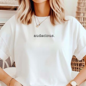 May include: White t-shirt with the word "audacious." printed in black on the front.