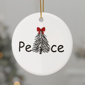 May include: A round white ceramic ornament featuring a black hand-drawn sketch of a pine tree topped with a small red bow. The word Peace is printed in black lettering with the tree replacing the letter a. A gold string is attached at the top.