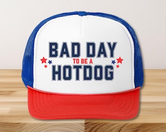 May include: A trucker hat with a white front panel, a red brim, and a blue mesh back. The hat features the text "BAD DAY TO BE A HOTDOG" in navy blue block letters, with red and blue star accents.