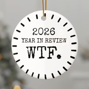 May include: A round white ceramic ornament featuring a black clock-like border. The text reads 2026 YEAR IN REVIEW WTF. The ornament is suspended by a thin gold string attached to a small hole at the top center of the circular white surface.