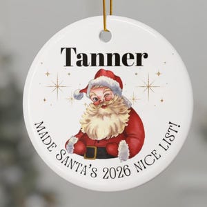 May include: A round white ceramic ornament featuring a winking Santa Claus illustration in the centre. The name Tanner is printed in black above Santa, and the text MADE SANTA'S 2026 NICE LIST! is printed in black along the bottom edge.