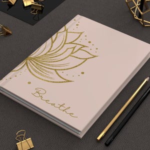 May include: A light pink journal with a gold floral design and the word "Breathe" in gold script. Two pencils, one gold and one black, are next to the journal. Gold binder clips are scattered around.