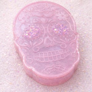 Puede incluir: Una caja de baratijas en forma de calavera rosa claro con diseños florales y de corazón. Los ojos están llenos de purpurina iridiscente. La caja está hecha de un material translúcido y se encuentra sobre una superficie rosa claro brillante.