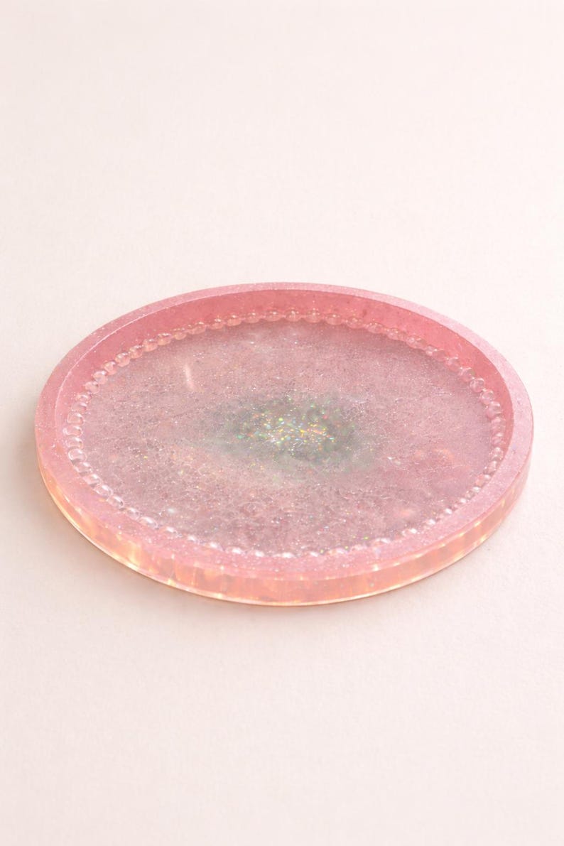 May include: A pink resin tray with a glittery, iridescent centre. The tray has a rounded oval shape with a beaded edge. The overall design is a soft, pastel aesthetic, suitable for decorative use.