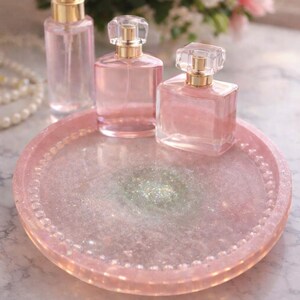 May include: A selection of pink perfume bottles with gold detailing on a pink glitter tray. The tray has a beaded edge. A spray bottle and two perfume bottles with crystal stoppers are visible. Soft pink flowers are in the background.