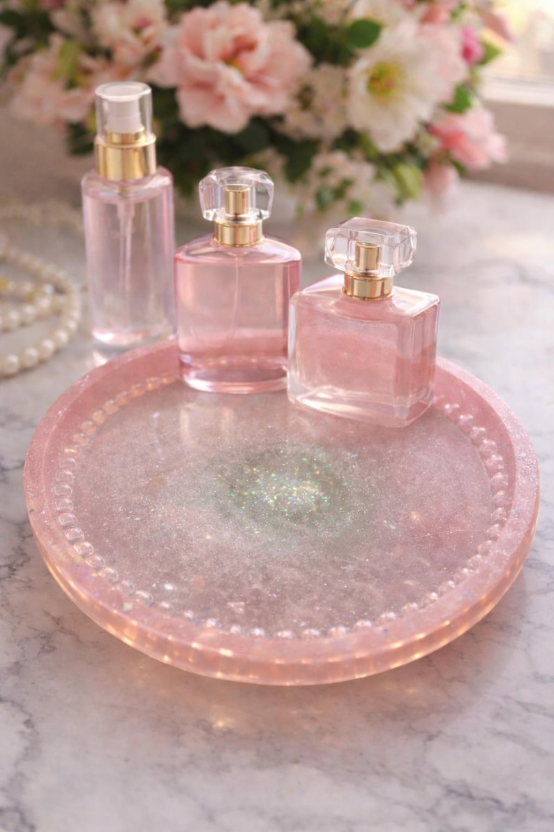 May include: A selection of pink perfume bottles with gold detailing on a pink glitter tray. The tray has a beaded edge. A spray bottle and two perfume bottles with crystal stoppers are visible. Soft pink flowers are in the background.