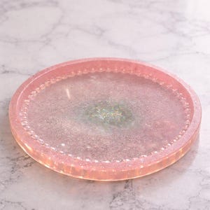 May include: A circular, pink resin tray with a glittery, iridescent centre. The tray has a raised edge with a beaded design. The overall aesthetic is delicate and decorative, suitable for serving or display.