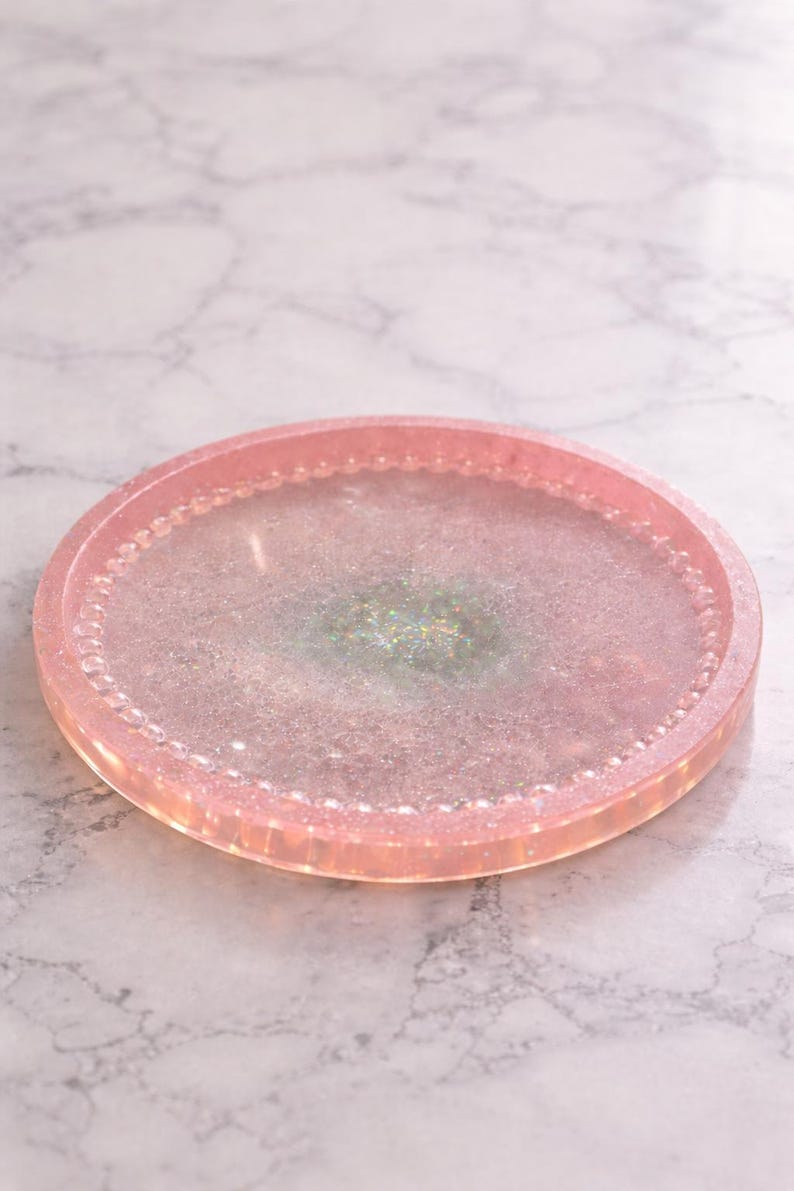 May include: A circular, pink resin tray with a glittery, iridescent centre. The tray has a raised edge with a beaded design. The overall aesthetic is delicate and decorative, suitable for serving or display.