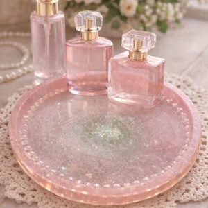 May include: Three pink perfume bottles with gold detailing are displayed on a matching pink tray with glitter. The tray sits on a white lace doily, with a blurred backdrop of pink flowers and pearls.