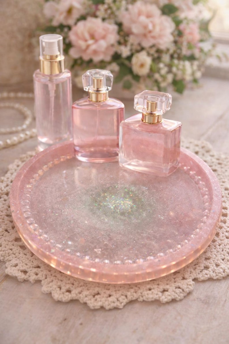 May include: Three pink perfume bottles with gold detailing are displayed on a matching pink tray with glitter. The tray sits on a white lace doily, with a blurred backdrop of pink flowers and pearls.