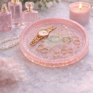 May include: A pink, glittery tray displays a gold watch, rings, and a necklace. Perfume bottles and a lit candle are in the background. The scene is soft and feminine, with a focus on jewellery and beauty products.