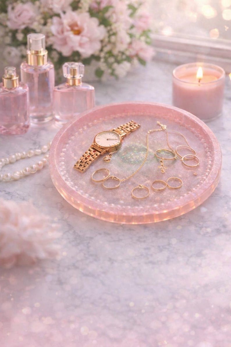 May include: A pink, glittery tray displays a gold watch, rings, and a necklace. Perfume bottles and a lit candle are in the background. The scene is soft and feminine, with a focus on jewellery and beauty products.