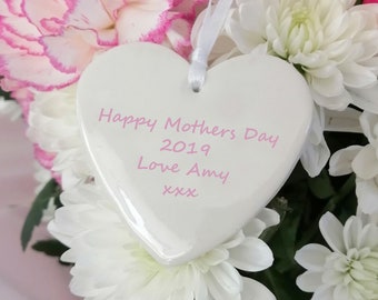 Puede incluir: Adorno de corazón blanco de cerámica con el texto "Happy Mother's Day 2019 Love Amy xxx" en letras rosas, rodeado de un ramo de flores blancas y rosas.