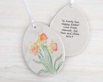 May include: Ceramic Easter egg ornament with a hand-painted design of yellow daffodils with orange centers. The back of the egg has a personalized message: "To Aunty Sue, Happy Easter! Love From Hannah, Sid, Tom and Chloe 2019".