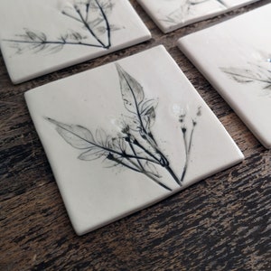 May include: A set of four square ceramic coasters. Each coaster has a botanical design of black and grey leaves and stems on a white background. The coasters are displayed on a dark wooden surface.