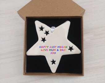 May include: A white ceramic star ornament with cut-out stars and the text "HAPPY 21ST MEGAN LOVE MUM & DAD XXX" in rainbow colours. The ornament is in a brown box with a black interior.