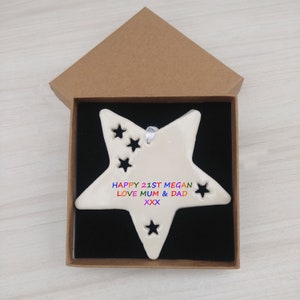 May include: A white ceramic star ornament with cut-out stars and the text "HAPPY 21ST MEGAN LOVE MUM & DAD XXX" in rainbow colours. The ornament is in a brown box with a black interior.