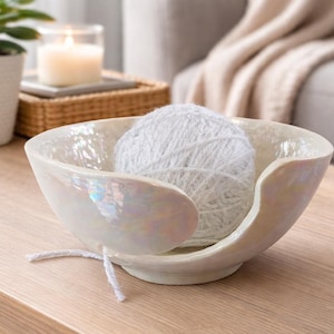 Iridescent ceramic yarn bowl with a pearlescent finish holding a white glitter yarn ball, styled on a light wooden table in a cosy living room setting with a soft blanket, candle, and plant in the background.