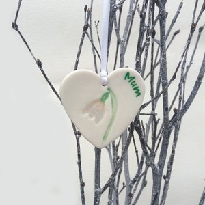 May include: White ceramic heart-shaped ornament with a green and white snowdrop flower and the word "Mum" written in green.