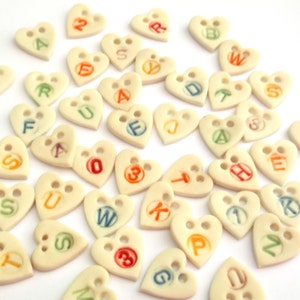 Puede incluir: Una colecci&oacute;n de botones en forma de coraz&oacute;n con letras y n&uacute;meros impresos en ellos. Los botones son blancos con letras de colores y est&aacute;n dispuestos sobre una superficie blanca.
