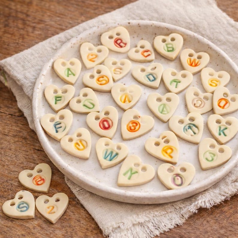 Personalised ceramic heart charms with colourful letters and numbers displayed on a neutral plate on a wooden surface.