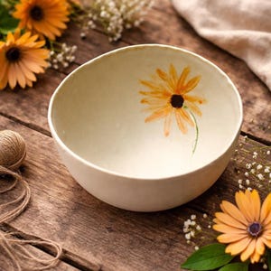 May include: Cream ceramic bowl featuring a hand-painted orange and black sunflower. The bowl sits on a rustic wooden surface, complemented by orange flowers and twine. A decorative item for the home, with a diameter of approximately 20 cm.
