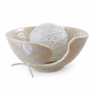 Iridescent ceramic yarn bowl with a pearly finish and side cut-out, holding a white glitter yarn ball, isolated on a clean white