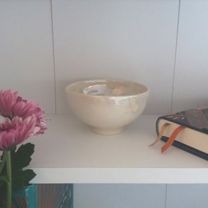 Puede incluir: Un cuenco de cer&aacute;mica blanca con un esmalte iridiscente. El cuenco est&aacute; sobre una estanter&iacute;a blanca con un libro y flores en el fondo.