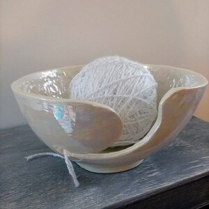 May include: A pearlescent white ceramic yarn bowl with a unique petal design, holding a ball of white yarn with silver threads. The bowl has a smooth, iridescent finish and a small opening for the yarn.