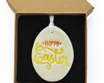 Puede incluir: Un adorno de cerámica blanca en forma de huevo con las palabras "Happy Easter" en letras amarillas y naranjas. El adorno está en una pequeña caja marrón.
