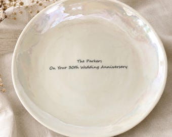May include: A round, white ceramic trinket dish with an iridescent, pearlescent finish. The center of the dish features black printed text that reads: The Parkers On Your 30th Wedding Anniversary. The surface has a slightly uneven, organic texture.
