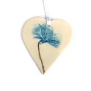 May include: A white ceramic heart-shaped ornament featuring a blue pressed flower design. The ornament has a white ribbon for hanging.