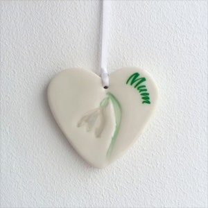May include: White ceramic heart-shaped ornament with a green and white snowdrop flower and the word "Mum" written in green.
