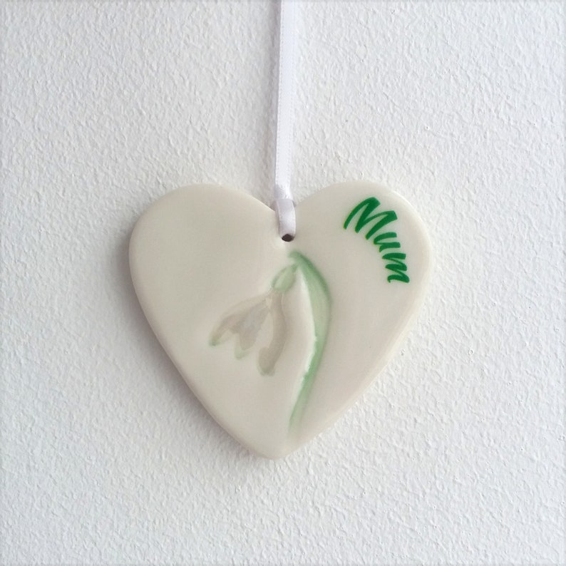 May include: White ceramic heart-shaped ornament with a green and white snowdrop flower and the word "Mum" written in green.