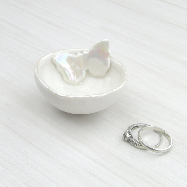 K&ouml;nnte beinhalten: Eine kleine, wei&szlig;e Keramik-Ringschale mit Schmetterlingsakzent. Die Schale ist rund mit einem leicht unregelm&auml;&szlig;igen Rand. Zwei silberne Ringe liegen neben der Schale. Der Schmetterling hat ein schillerndes Finish.