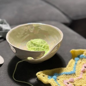 May include: A ceramic yarn bowl with a light green interior and a curved opening. A ball of bright green yarn sits inside the bowl, with the yarn extending through the opening. A yellow and blue crocheted item is in the foreground.