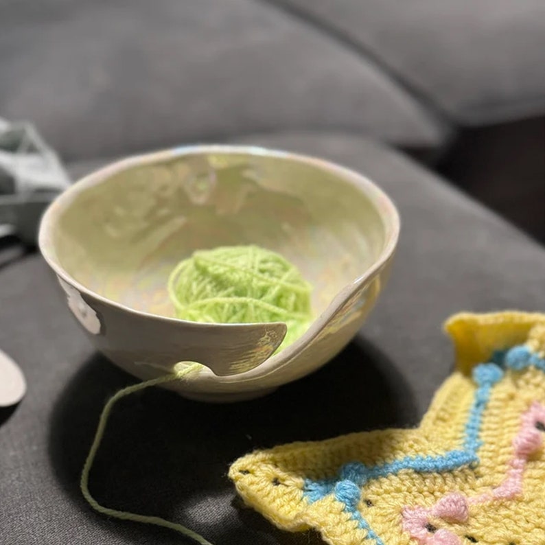 May include: A ceramic yarn bowl with a light green interior and a curved opening. A ball of bright green yarn sits inside the bowl, with the yarn extending through the opening. A yellow and blue crocheted item is in the foreground.