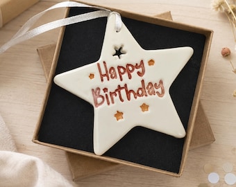 May include: A white ceramic star-shaped ornament featuring the brown text Happy Birthday and small brown star accents. The ornament includes a white ribbon for hanging and is presented inside a square brown gift box with a black insert.