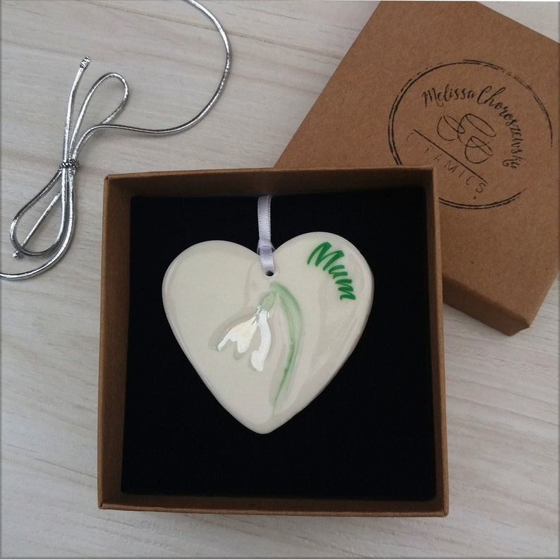 May include: White ceramic heart-shaped ornament with a green snowdrop flower and the word "Mum" painted on it. The ornament is in a brown box with a black velvet lining.