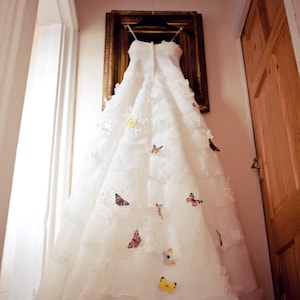 A beautiful ruffled silk wedding dress hangs on a picture frame at the top of a flight of stairs. there are silk butterflies pinned to the skirt.