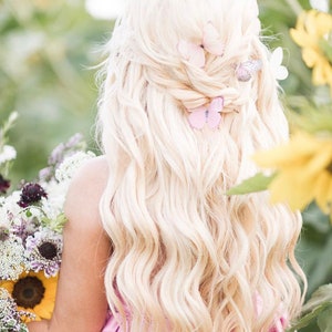 A lady with wavy long blond hair is holding a bunch of flowers including sunflowers. In the back of her hair are three pretty pale pin butterfly hair clips