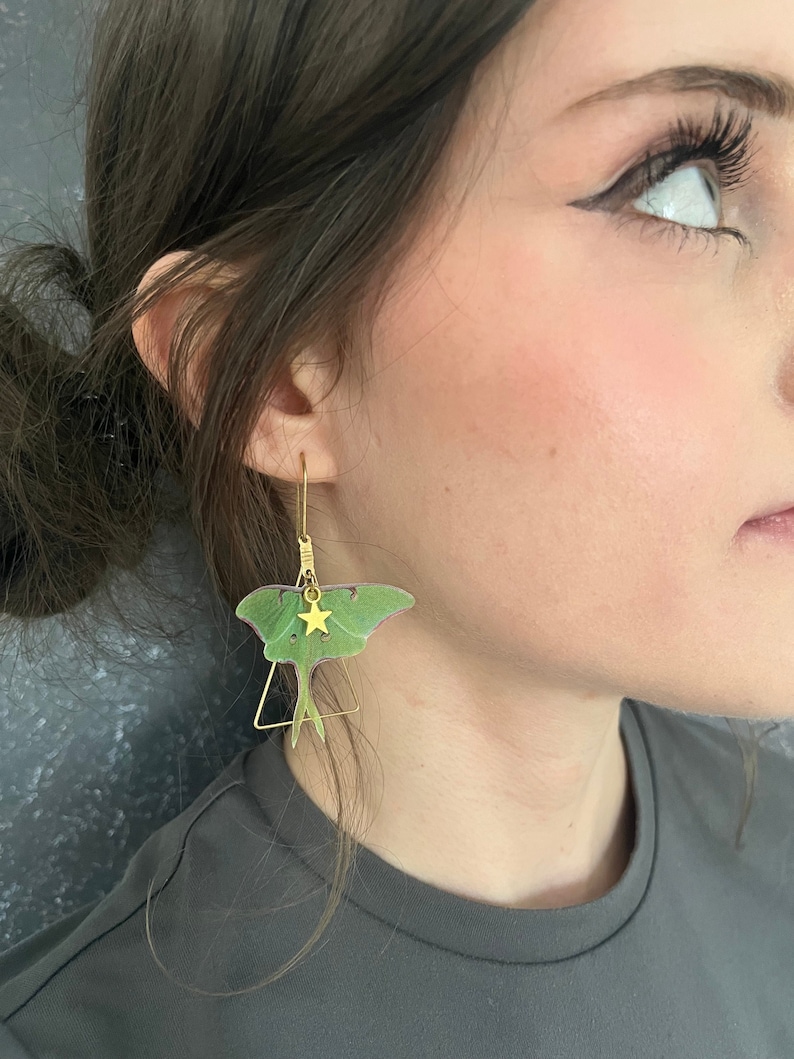 a girl with black wing eyeliner wears brass triangle earrings with a delicate green silk moth  and a gold star charm.