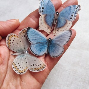 A hand holds three palest blue silk butterflies. Each little butterfly has a tiny sparkling crystal on its head. these silk butterflies are slipped onto bobby pins and are the perfect wedding accessory.